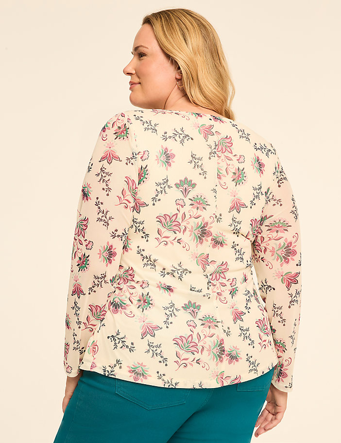 A woman in a floral blouse and teal pants stands against a plain, light-colored background.