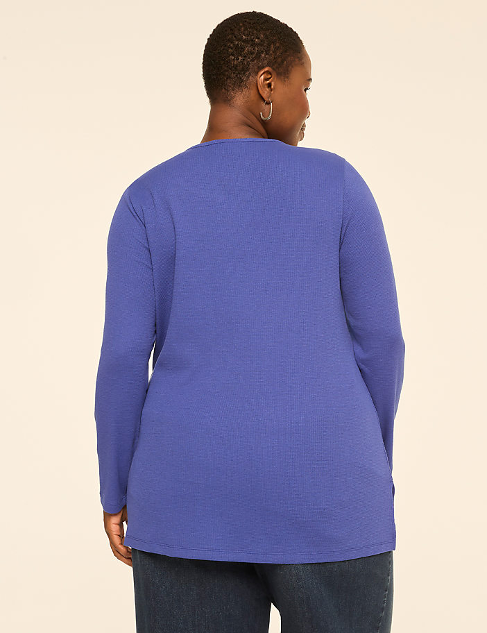 A woman in a blue long-sleeved shirt and jeans stands against a plain, light-colored background.