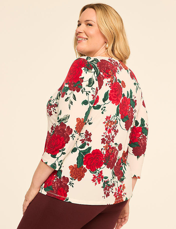 A woman in a floral top and maroon pants smiles over her shoulder against a plain background.