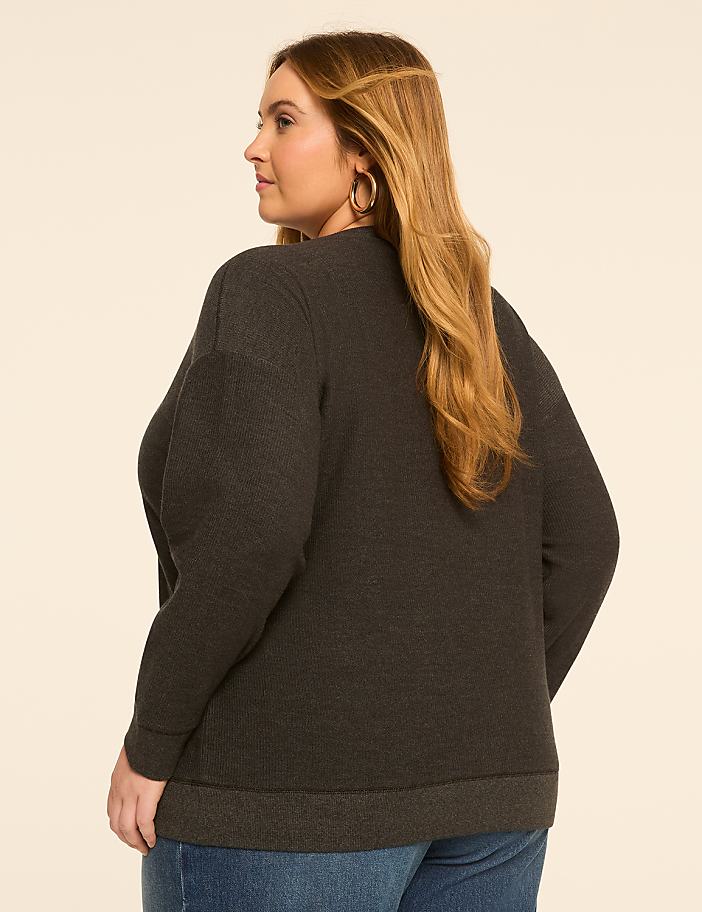 A woman in a dark gray sweater and blue jeans stands against a plain, light-colored background.