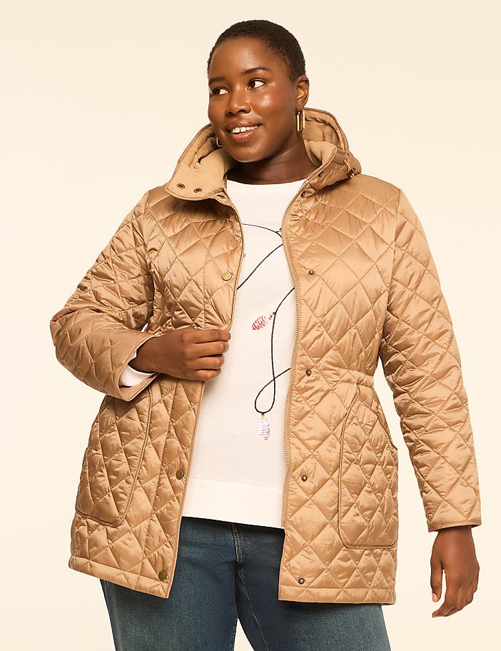A woman in a tan quilted jacket and white sweater smiles against a plain, light-colored background.