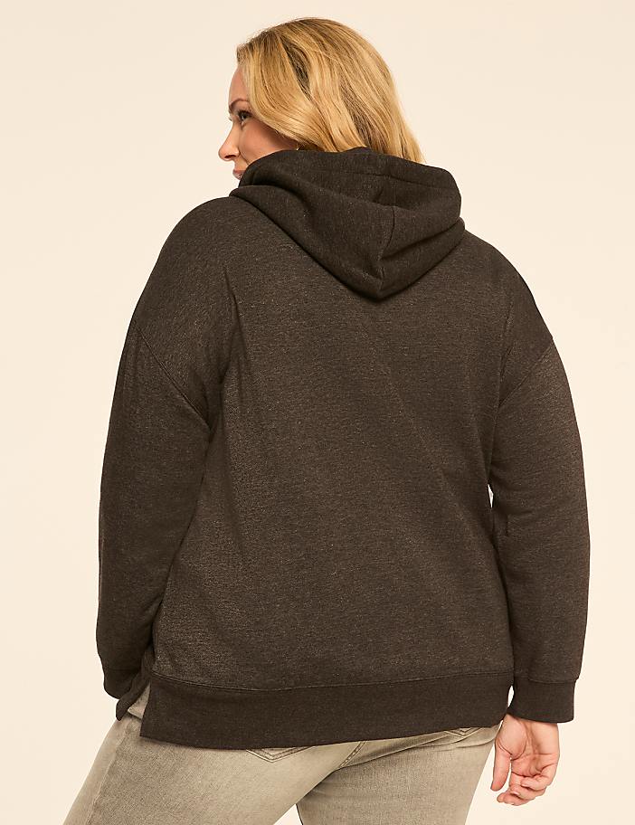 A woman in a dark grey hoodie and light grey jeans stands against a plain, light beige background.