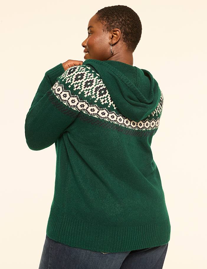 A woman in a green hooded sweater with a fair isle pattern on the back stands against a plain, light-colored background.