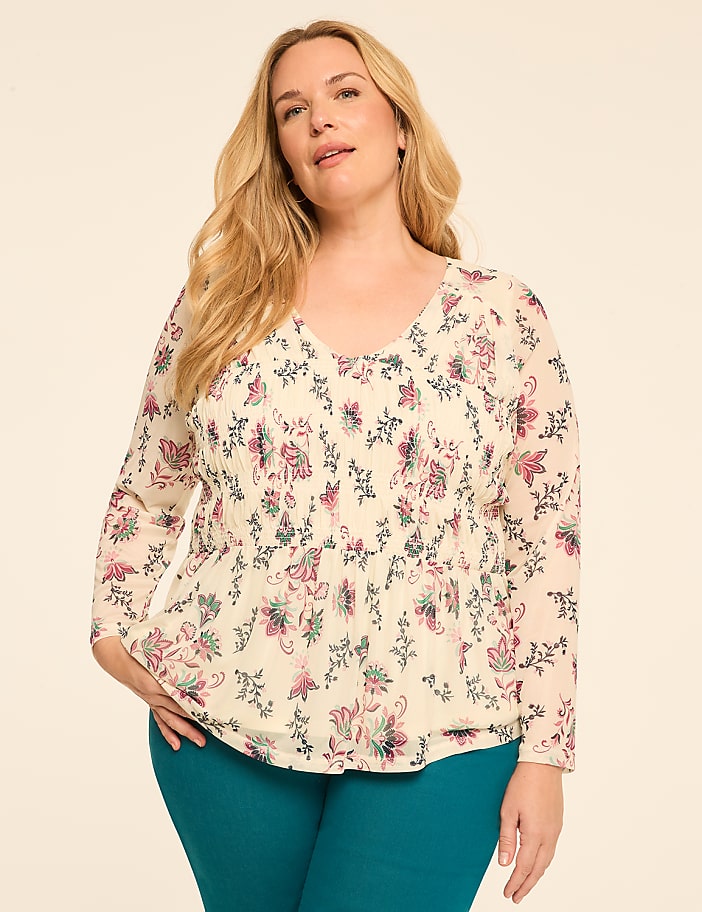 A woman with blonde hair and fair skin, wearing a cream-colored floral blouse and teal pants, stands against a plain beige background.