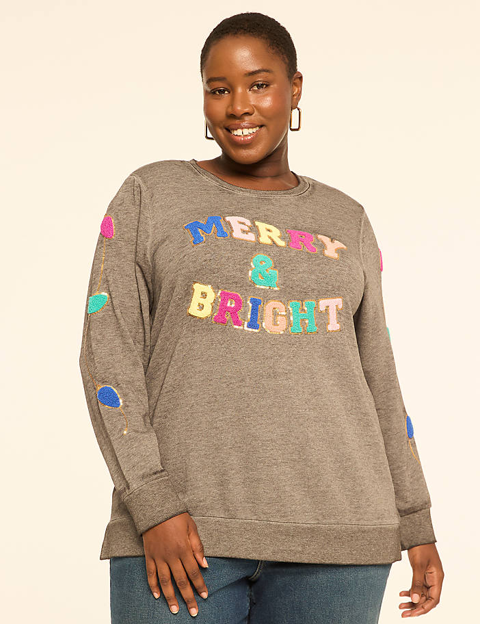A woman in a gray "Merry & Bright" sweatshirt stands against a plain, light-colored background.