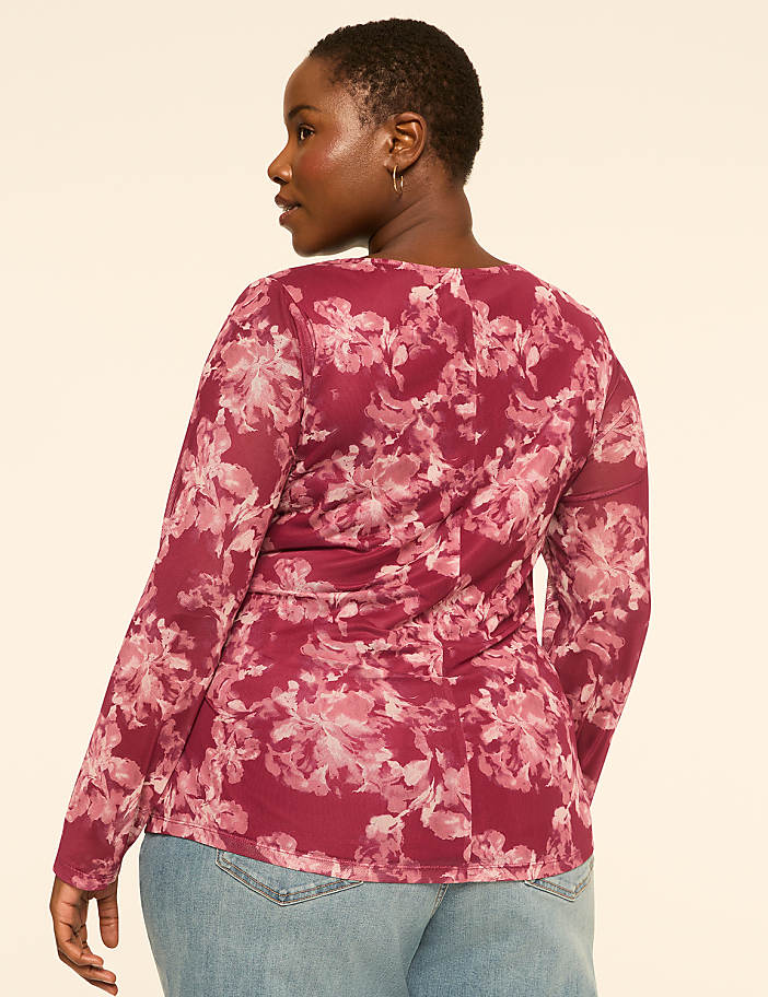 A woman in a floral mesh top and jeans stands against a plain, light-colored background.