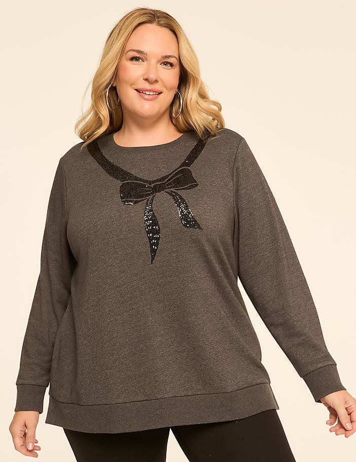 A woman in a dark grey sweatshirt with a sequined bow neckline stands against a plain, light beige background.