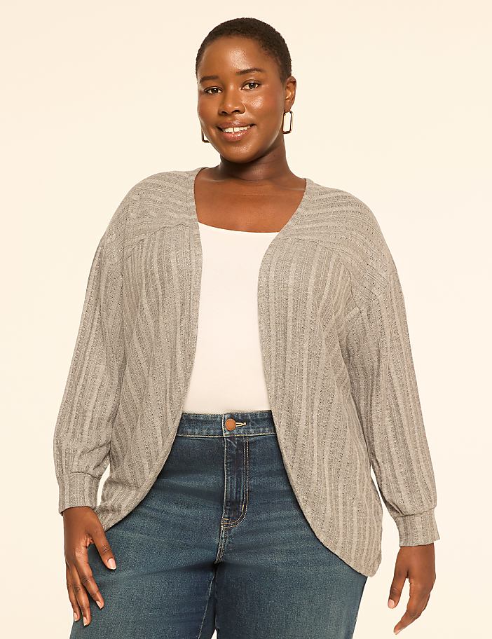 A smiling woman in a gray cardigan and blue jeans stands against a plain, light-colored background.
