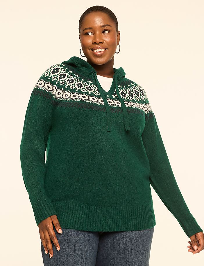 A smiling woman in a green hooded sweater with a fair isle pattern stands against a plain, light-colored background.