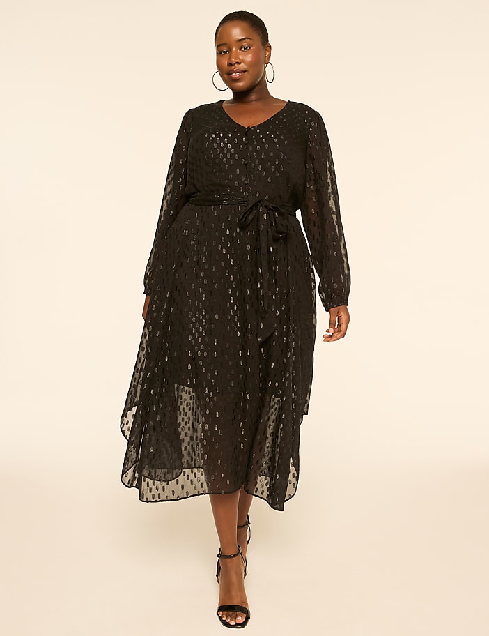 A woman in a black patterned dress stands against a plain, light-colored background.