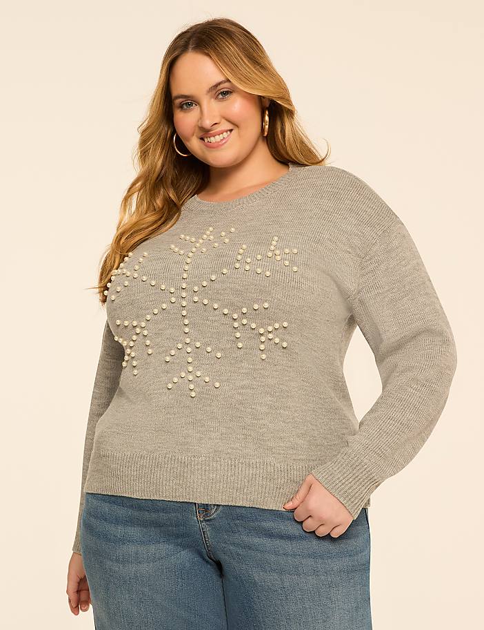 A smiling woman in a pearl-adorned snowflake sweater and jeans stands against a plain, light-colored background.