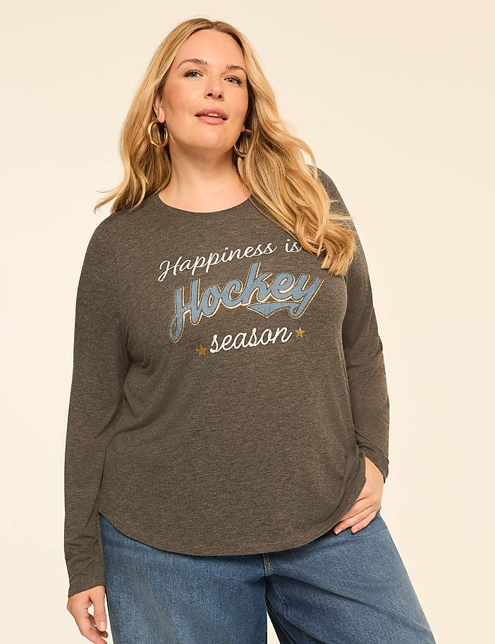 A woman in a long-sleeved shirt with "Happiness is Hockey season" printed on it stands against a plain, light-colored background.