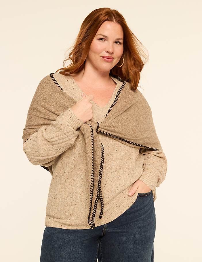 A woman with auburn hair poses in a beige sweater and jeans, with a tan shawl draped over her shoulders, against a plain, light-colored background.