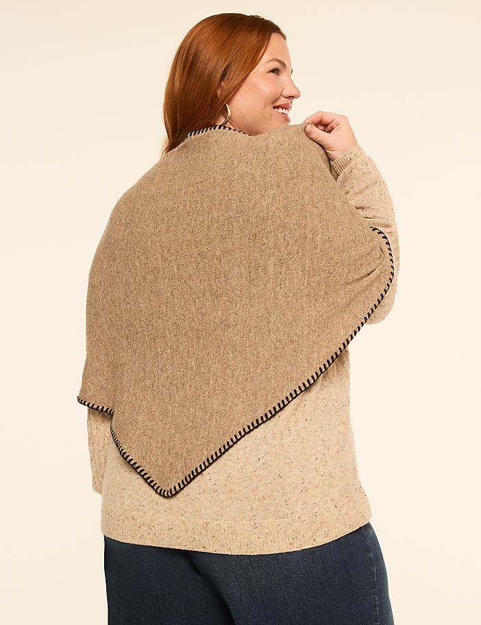 A woman with auburn hair models a tan poncho with black trim over a speckled tan sweater and blue jeans against a plain cream background.