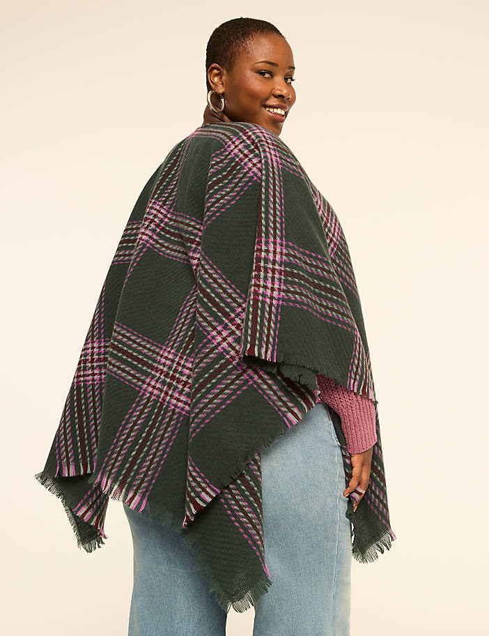 A woman in a green and purple plaid poncho and pink sweater smiles over her shoulder against a plain, light background.