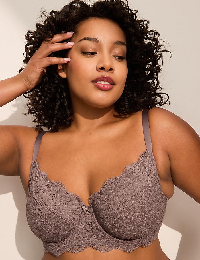 A woman with curly dark hair poses in a taupe lace bra against a plain, light-colored background.