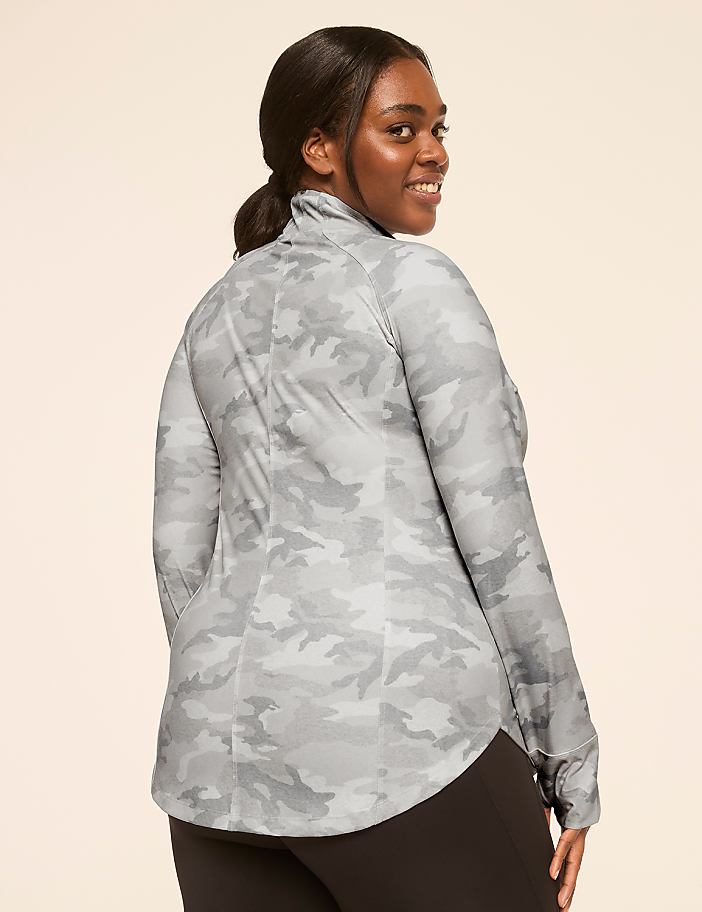A woman in a camouflage top and black leggings smiles over her shoulder against a plain, light background.