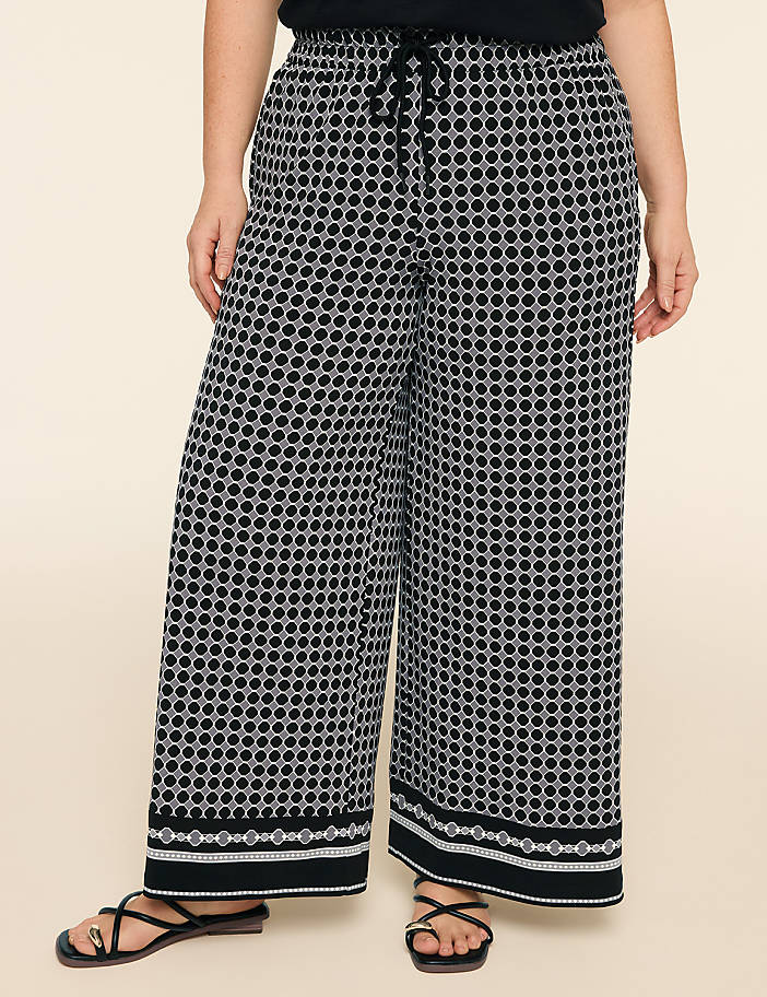 A person wearing black and white patterned wide-leg pants with a decorative hem stands against a plain beige background.