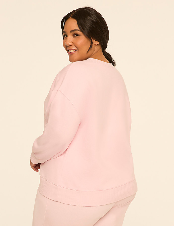 A woman in a pink sweatshirt and pants smiles over her shoulder against a plain, light background.