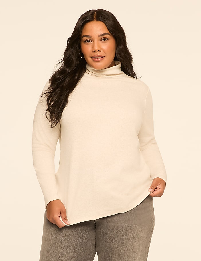 A woman in a cream turtleneck sweater and gray pants stands against a plain, light beige background.