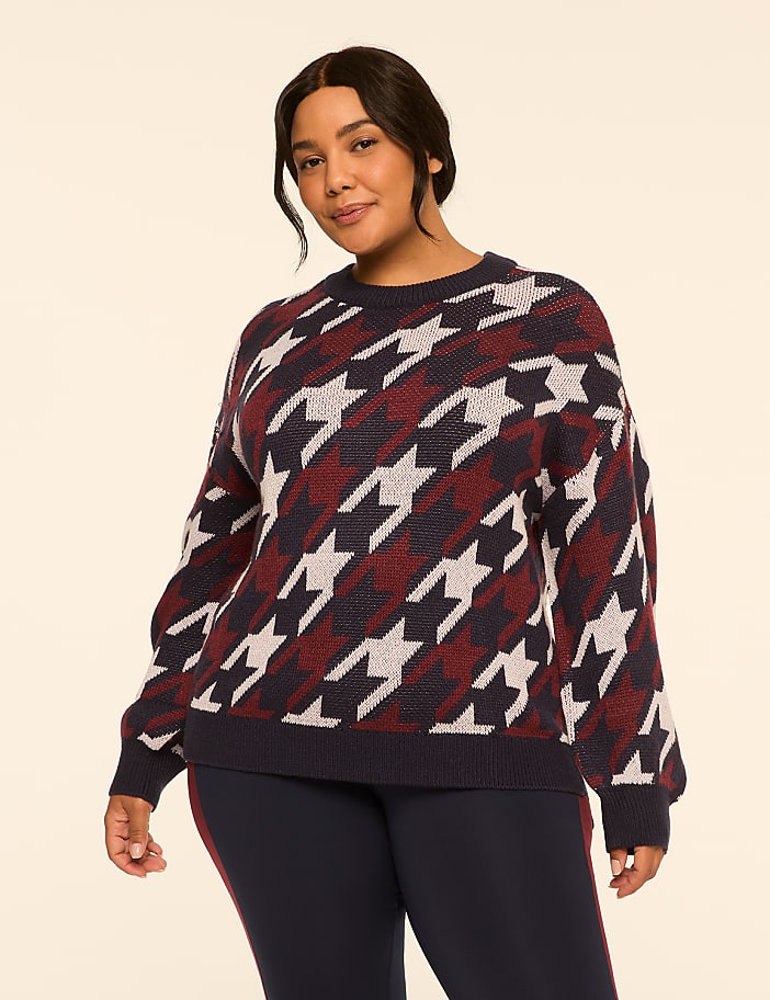 A woman in a houndstooth sweater and dark pants stands against a plain, light-colored background.