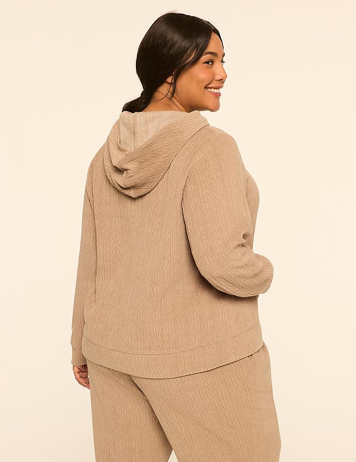 A woman in a tan hoodie and pants stands against a plain, light background.