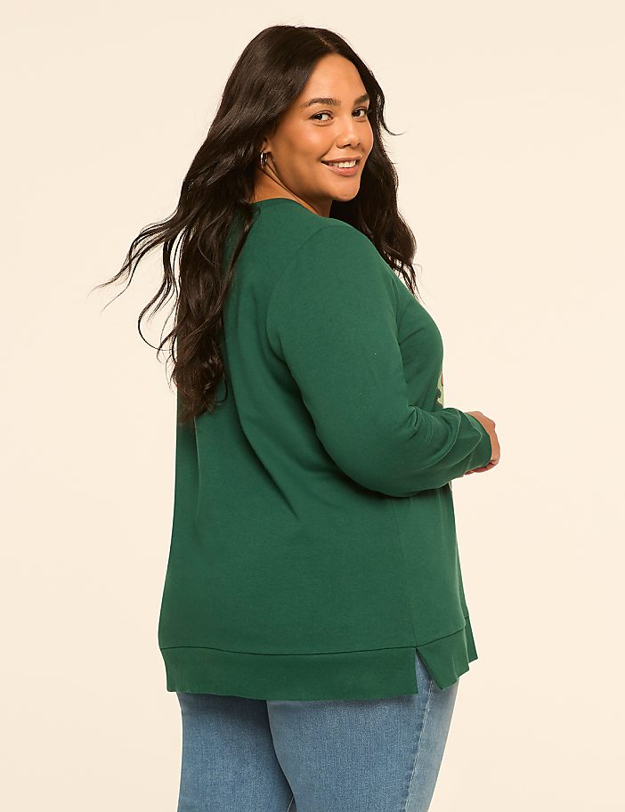 A woman in a green sweatshirt and blue jeans smiles over her shoulder against a plain, light-colored background.
