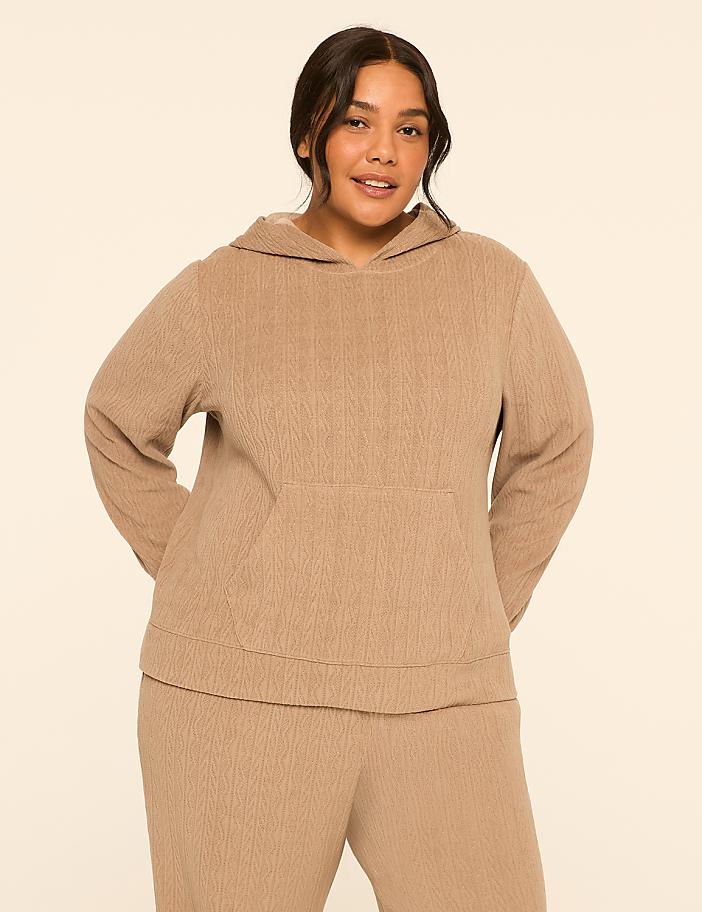 A woman in a tan hoodie and matching pants stands against a plain, light tan background.
