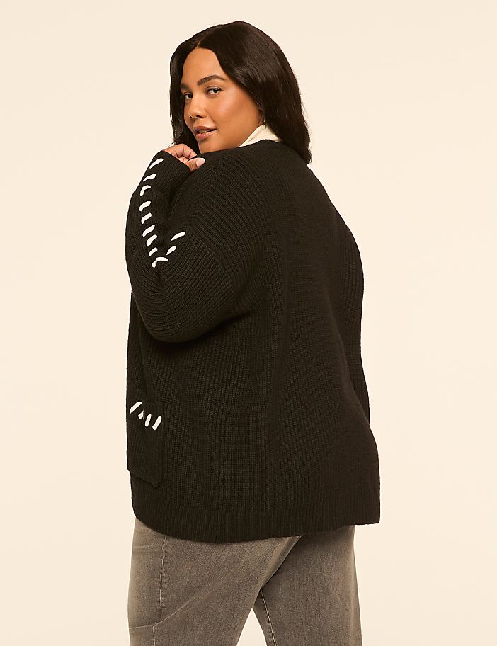 A woman in a black cardigan with white stitching on the sleeves and grey jeans stands against a plain, light-colored background.