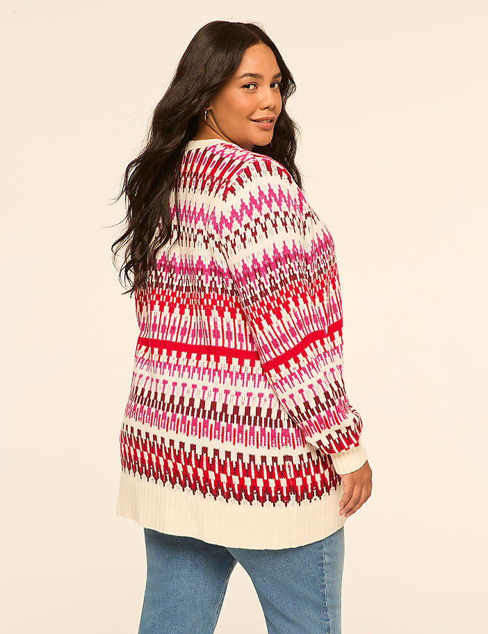 A woman in a colorful patterned sweater and jeans stands against a plain, light-colored background.