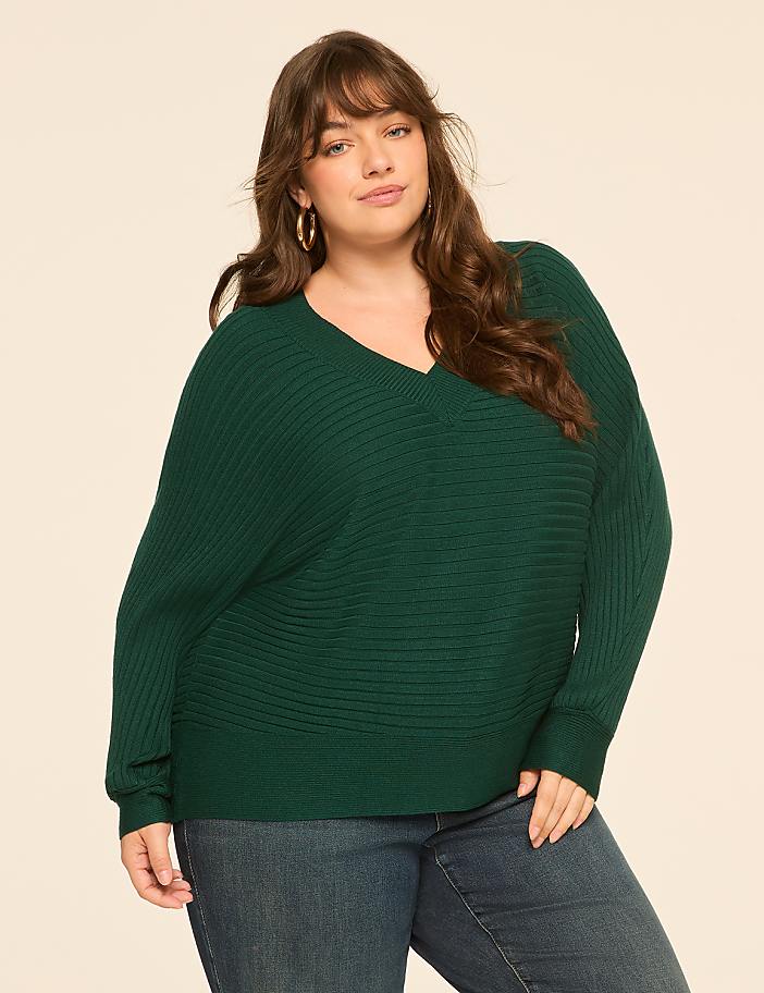 A woman in a green sweater and jeans stands against a plain, light-colored background.