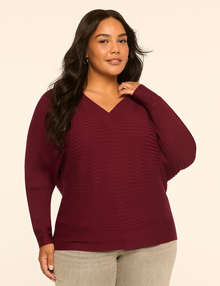 A woman in a maroon v-neck sweater and grey pants stands against a plain, light-colored background.