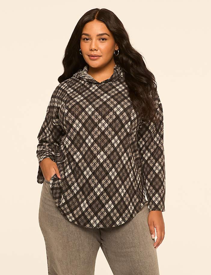 A woman with long, dark, wavy hair stands in the foreground wearing a gray and black plaid hoodie and gray jeans, against a plain, light beige background.