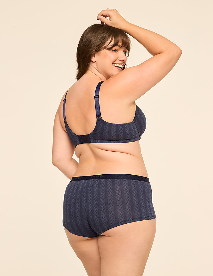 A woman in a navy blue patterned bra and matching underwear poses with her arm raised against a plain, light-colored background.