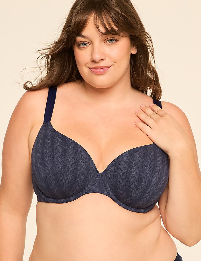 A woman with a hand on her chest wears a patterned blue bra against a plain, light-colored background.