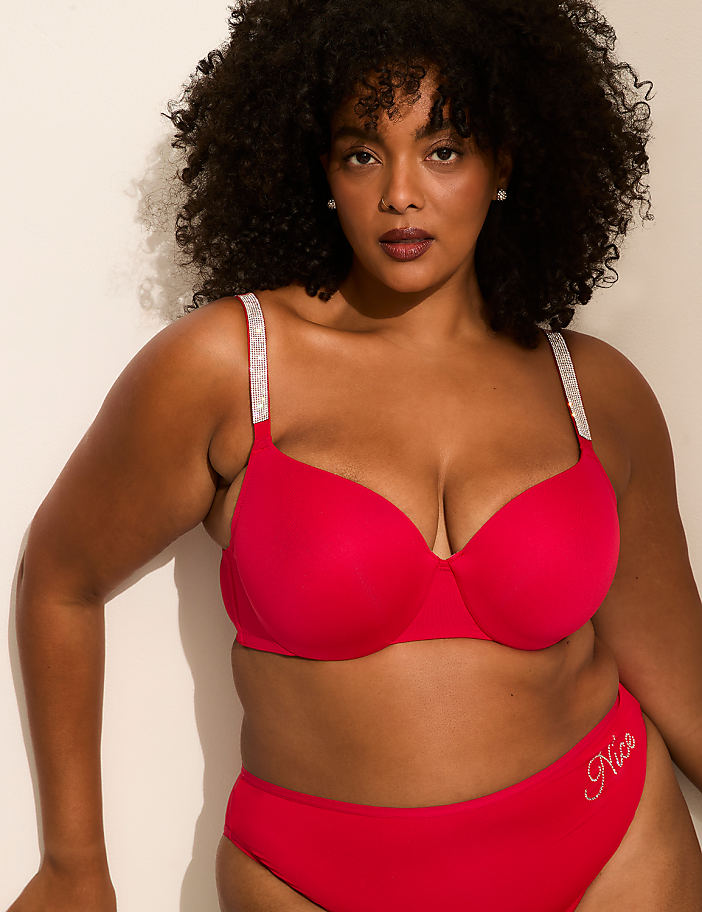A woman with curly hair poses in a red bra with jeweled straps and matching underwear against a plain white background.