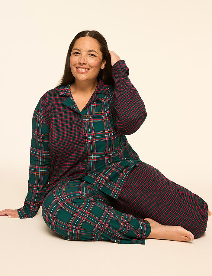 A smiling woman in plaid pajamas sits on a light-colored floor against a plain, light background.
