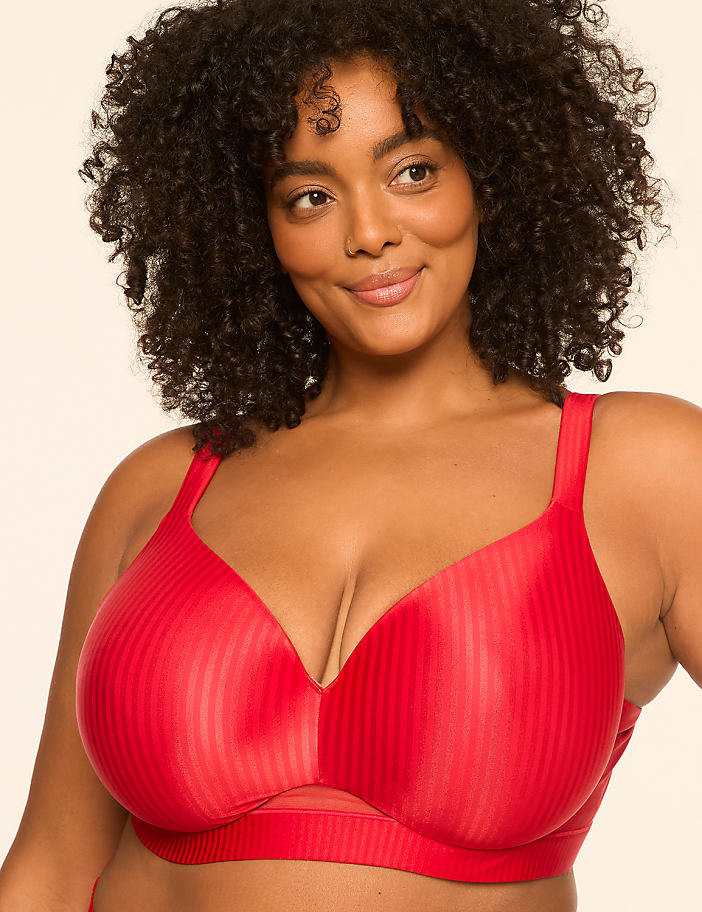 A woman with curly dark hair smiles gently, wearing a red ribbed bra against a soft, peachy background.