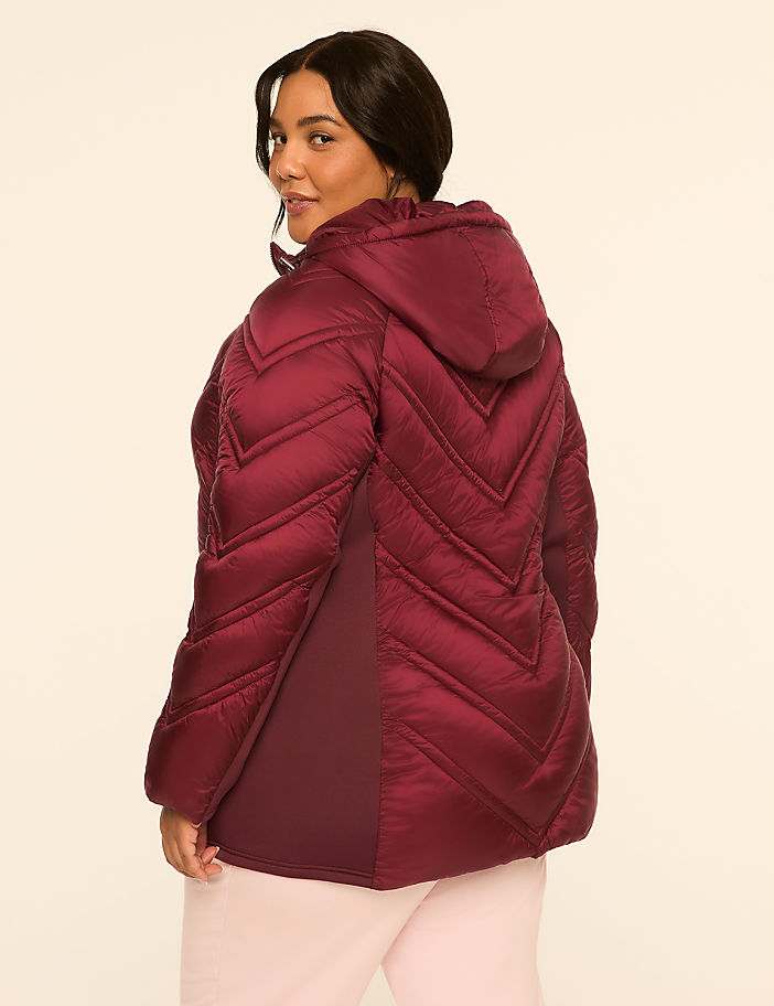 A woman in a burgundy puffer jacket with chevron stitching stands against a plain, light-colored background.