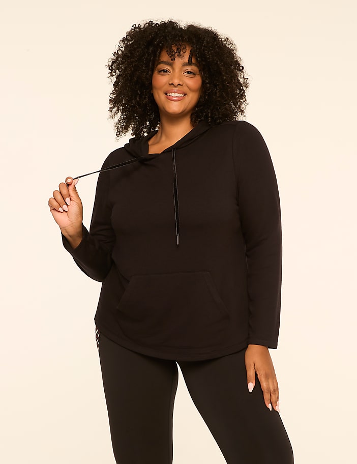 A smiling woman in a black hoodie and leggings stands against a plain, light-colored background.