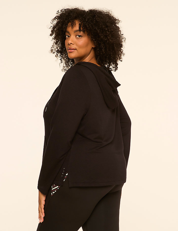 A woman with curly dark hair, wearing a black hooded sweatshirt and black pants, stands against a plain, light-colored background.