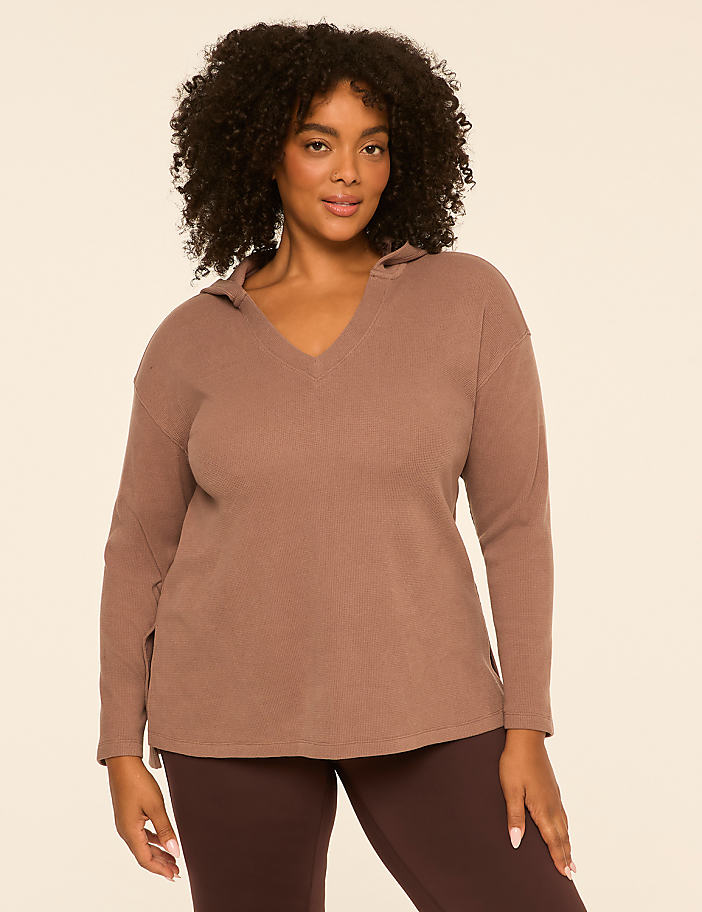 A woman in a brown hoodie and leggings stands against a plain, light-colored background.