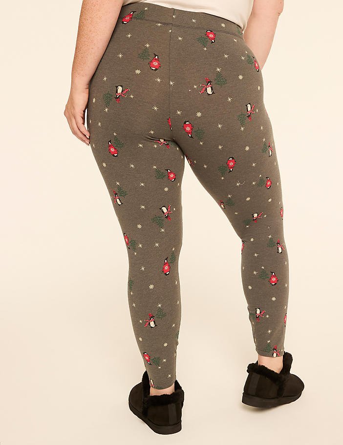 A person wearing grey leggings with a festive pattern of robins and stars stands on a plain cream background, with black slippers on their feet.