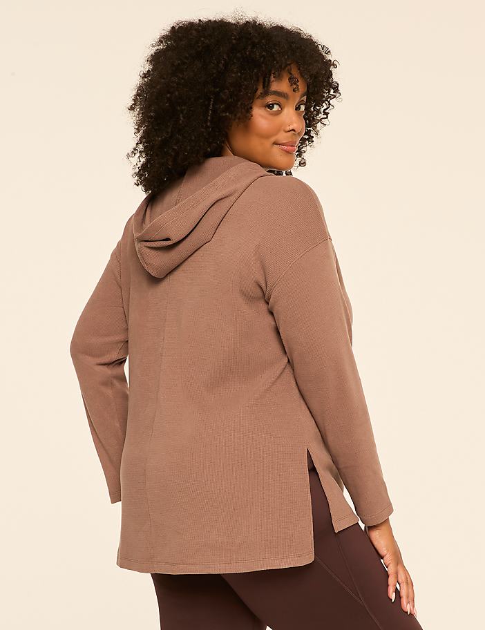 A woman in a brown waffle-knit hoodie and dark leggings poses against a plain beige background.