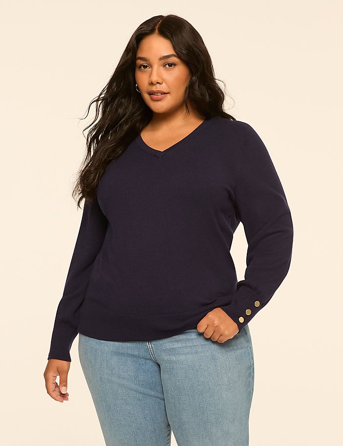 A plus-size woman in a navy v-neck sweater and light blue jeans stands against a plain, light beige background.