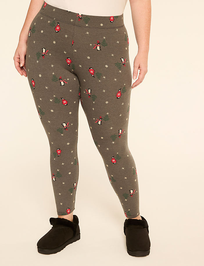 A person wearing grey leggings with a festive bird and snowflake pattern stands on a plain background, with black fuzzy slippers on their feet.