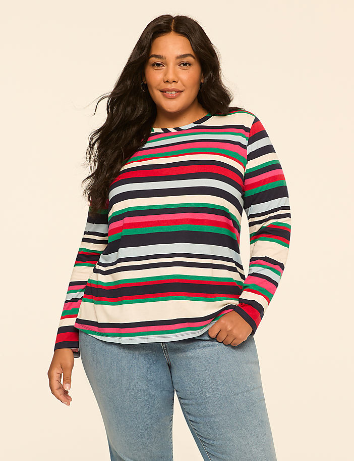 A woman with dark hair and a striped long-sleeved shirt stands against a plain, light-colored background.