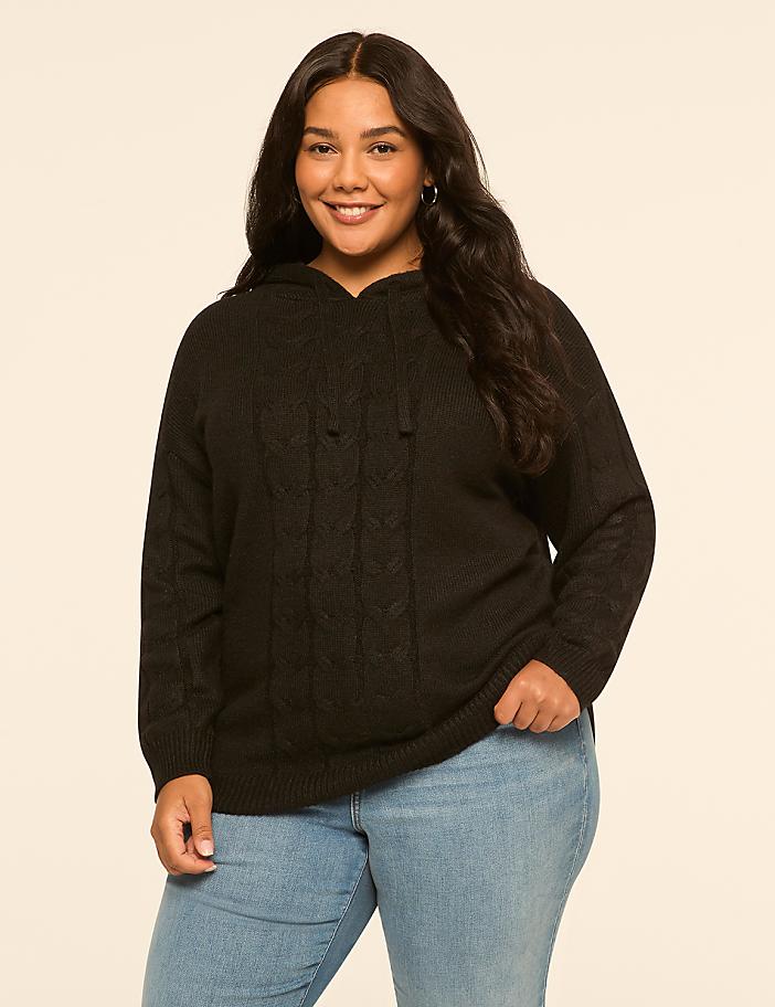 A smiling woman in a black cable-knit hoodie and light blue jeans stands against a plain, light beige background.