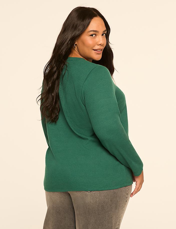 A woman in a green sweater and gray pants poses against a plain, light-colored background.