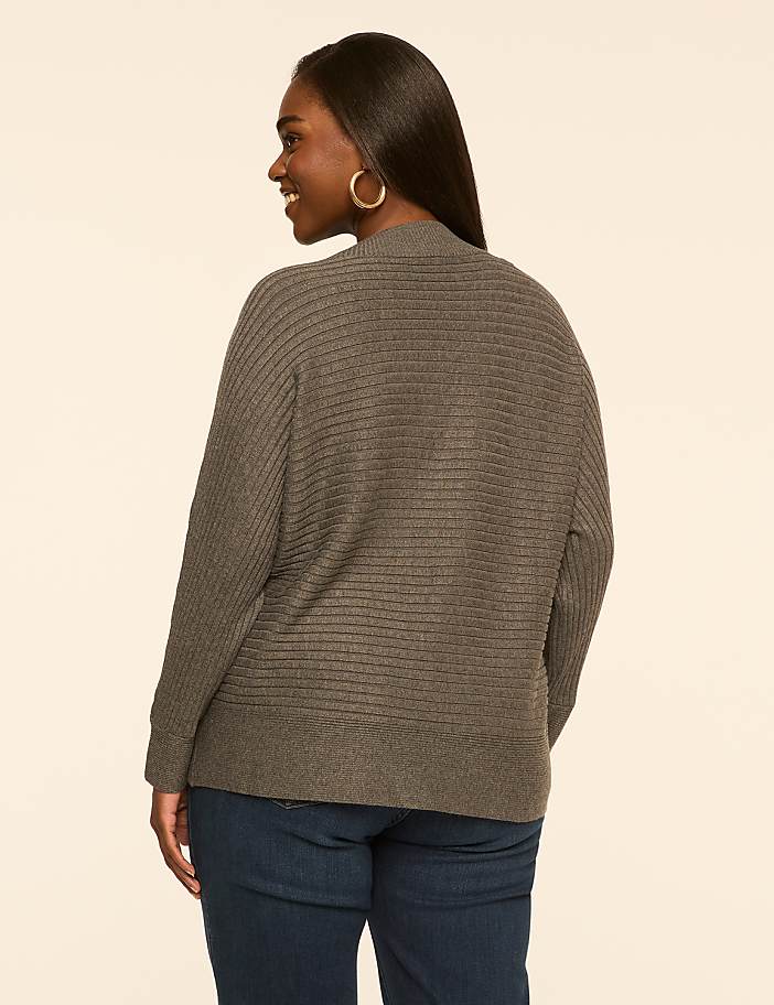 A woman in a gray cardigan and blue jeans stands against a plain, light brown background.